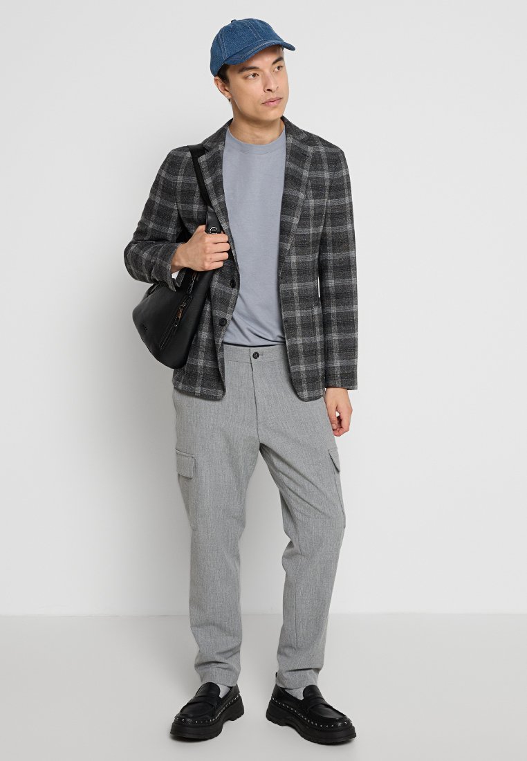 Grauer Karoblazer, hellgraues T-Shirt, graue Cargo-Hosen mit Taschen, schwarze Schuhe und blaue Mütze; lässiger, aber taillierter Look.