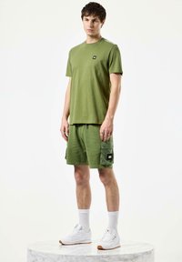 Grøn bomulds t-shirt og matchende shorts med cargo-lommer. Hvide sneakers med gummisul og ribbede hvide strømper. Minimalistiske designelementer.