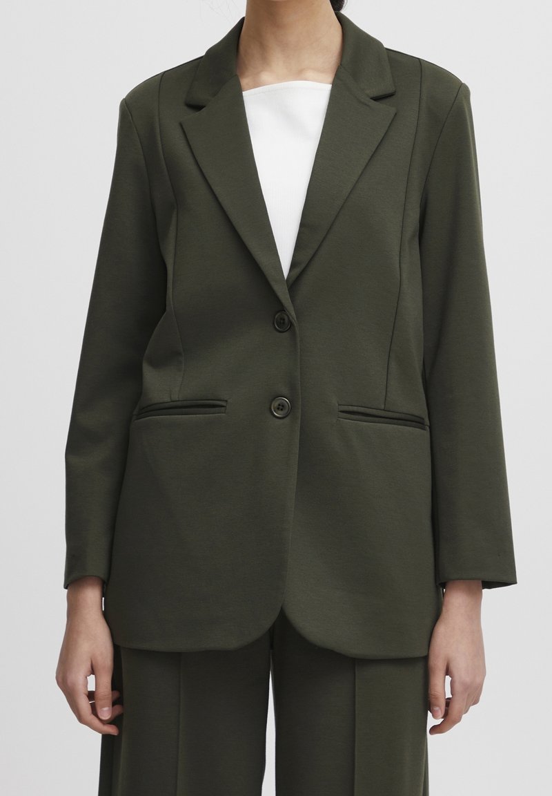 Femme portant un blazer vert olive ajusté avec deux boutons et un pantalon assorti sur un top blanc, debout devant un fond uni.