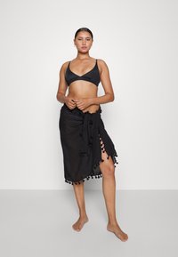 Top de biquini preto com uma saia preta de estilo envolvente, apresentada com franjas ao longo da bainha, nós circulares e uma fenda lateral. Textura de tecido suave.