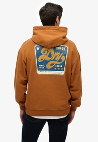 Sudadera marrón con un gran gráfico azul y amarillo en la parte posterior, que presenta el texto "Super Dry" y estrellas. Textura suave con puños de canalé.