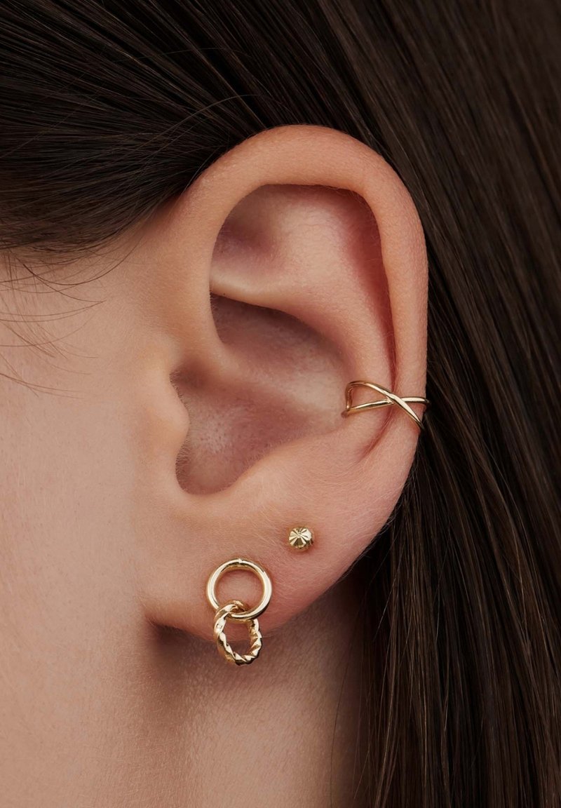 Goldene Creolen mit gedrehter Gestaltung, einem goldenen Stecker und einem Ohrcuff in Kreuzform, vor braunem Haar.