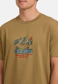 Zielona oliwkowa bawełniana koszulka z graficznym wzorem przedstawiającym góry, fale i palmy, z napisem "adventure division billabong" na czerwono.