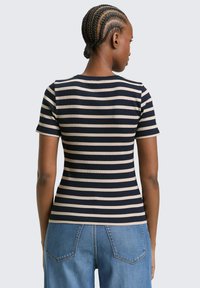 Koszula w paski z krótkim rękawem w kolorze granatowym i beżowym, wykonana z prążkowanego materiału, z okrągłym dekoltem i dopasowanym krojem. Noszona z wysokim stanem dżinsów.