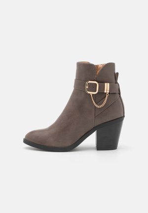 Bottines en daim marron avec un talon carré, dotées d'une sangle latérale et de matériaux métalliques dorés, incluant une boucle de ceinture et un détail en chaîne.