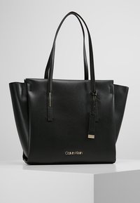 Mustat nahkainen Calvin Klein -käsilaukku kaksoisolkaintien ja kultalogon kanssa, esillä valkoisella jalustalla harmaata taustaa vasten.