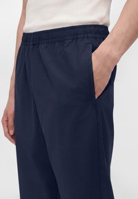 Homme portant un pantalon bleu marine à taille élastique avec la main dans la poche latérale, montré de la taille jusqu'au milieu de la cuisse sur un fond uni.