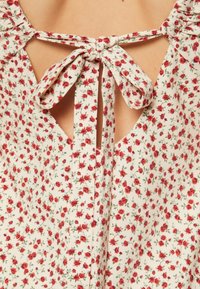 Tejido blanco con pequeño patrón floral rojo, con una apertura en la parte posterior con un ojal atado con tiras de tela. Escote texturizado con pliegues.