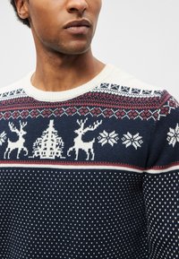 Tmavomodrý pletený sveter s bielymi a červenými vzormi, obsahujúci soby, vianočný stromček a snehové vločky. Výstrih dookola krku a textúrovaná tkanina.
