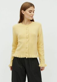 Sárga, puha gyapjú kardigán fodros szélekkel, kerek nyakkivágással és gombos elülső résszel. Hosszú ujjú, fodros mandzsettájú.