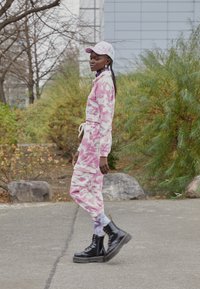 Macacão camouflage rosa com cintura de cordão, mangas longas e bolsos laterais; combinado com botas pretas de atacadores e um boné correspondente.