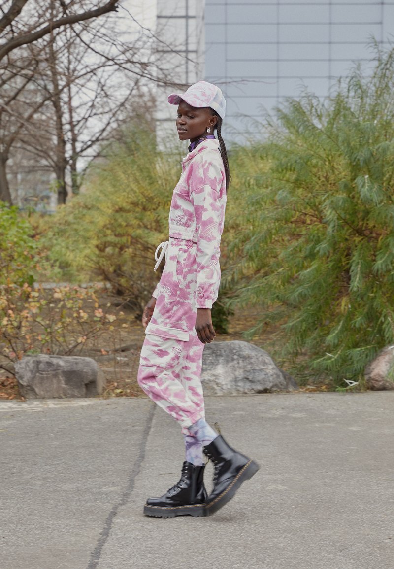 Macacão camouflage rosa com cintura de cordão, mangas longas e bolsos laterais; combinado com botas pretas de atacadores e um boné correspondente.