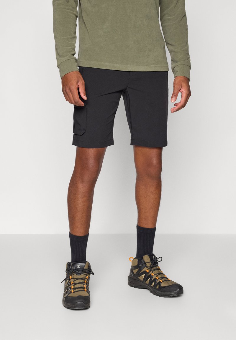 Schwarze Cargo-Shorts mit flachen Nähten und Seitentaschen. Kombiniert mit dunklen Socken und robusten, mehrfarbigen Wanderschuhen. Olivgrünes Langarmoberteil.