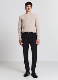 Beiger Strickpullover mit Rundhalsausschnitt, kombiniert mit schwarzen, plissierten Hosen und schwarzen Stiefeletten, vor neutralem Hintergrund.