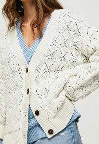 Bílý pletený kardigan s texturovaným krajkovým vzorem, s žebrovaným lemem a manžetami a velkými hnědými knoflíky. Pod ním modrý svetřík s výstřihem do V.