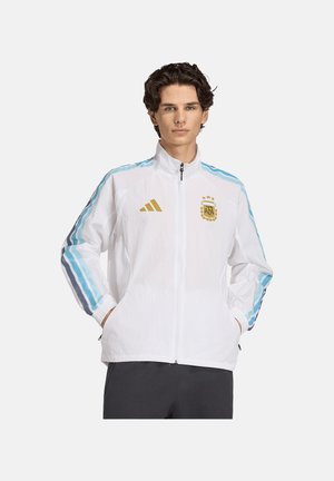 Młody mężczyzna w białej kurtce na zamek z niebieskimi paskami na rękawach i złotymi logo Adidas i AFA na piersi, ręce w kieszeniach.