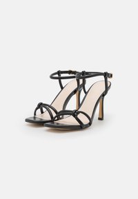 Sandalias de tacón de cuero negro con tiras delgadas y hebilla en el tobillo. Presenta detalles anudados en la parte frontal y una plantilla suave de color beige.