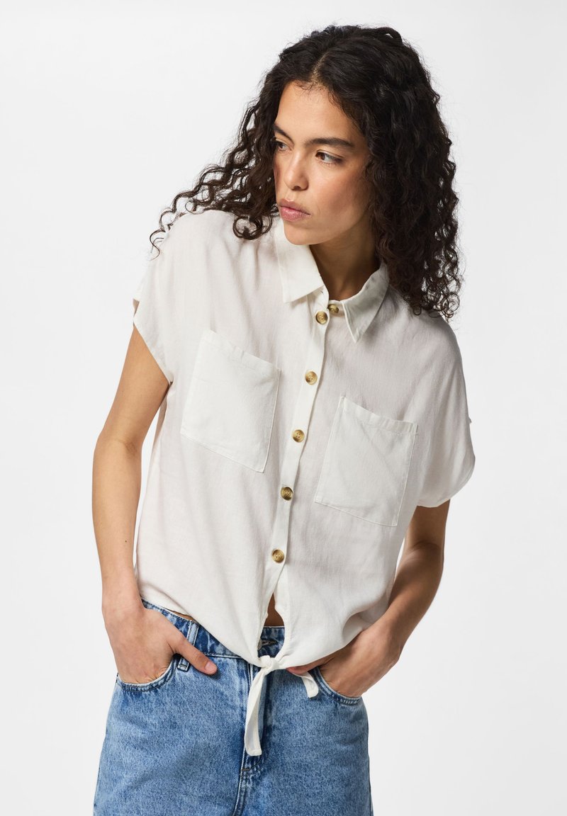 Femme aux cheveux bouclés portant une chemise blanche à manches courtes nouée à la taille et un jean bleu, mains dans les poches, regardant sur le côté.