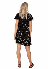 Robe noire à motif floral blanc, manches courtes, taille cintrée. Le tissu semble léger et tombe jusqu'à mi-cuisse. Sandales plates visibles.