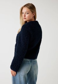 Kobieta z długimi blond włosami, ubrana w ciemnoniebieski sweter o fakturze i jasnoniebieskie dżinsy, patrząca przez ramię na tle jednolitego tła.
