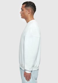 Sweatshirt blanc oversize avec un col rond, des épaules tombantes et des poignets côtelés. Texture douce et détails sur les coutures latérales sont visibles.
