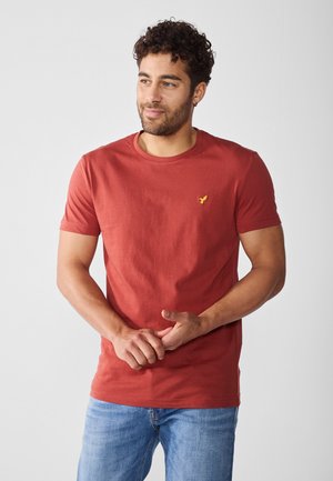 T-shirt rossa a girocollo in cotone, con maniche corte e un piccolo logo giallo sul petto, abbinata a jeans azzurri chiari.