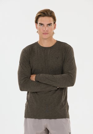 Jeune homme aux cheveux châtain clair portant une chemise à manches longues gris foncé et un pantalon gris clair, bras croisés devant un fond blanc uni.