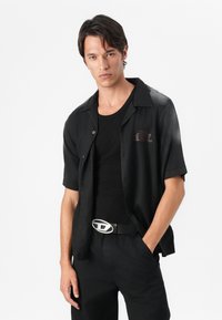 Black short-sleeve shirt with a subtle logo, worn over a black fitted shirt and paired with black trousers and a silver oval belt buckle.