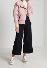 Veste matelassée rose clair avec col en fausse fourrure, poches zippées, associée à un jean ample foncé et des baskets grises. Texture lisse et coupe décontractée.