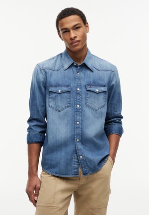 Mladý muž v modrej rifľovej košeli s gombíkmi a vyhrnutými rukávmi a béžových cargo nohaviciach, stojaci s jednou rukou v vrecku.