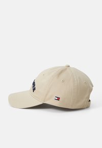 Beige bomullcaps med en buet skygge, brodert tekst på fronten, og en liten merke-logo-patch på siden. Justerbar stropp bak.