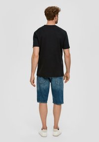 Černé tričko s krátkým rukávem, spárované s modrými džíny po kolena. Model nosí bílé tenisky. Jednoduchý, ležérní outfit bez vzorů.