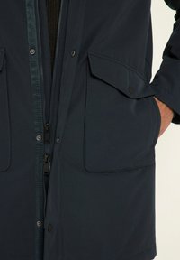 Parka bleu marine avec double fermeture éclair et boutons-pression, grandes poches avant et texture lisse. Convient à diverses conditions météorologiques.