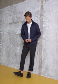 Navy zip-up jacket with a collar, white t-shirt underneath, black tapered trousers, and black lace-up shoes, set against a textured gray wall.