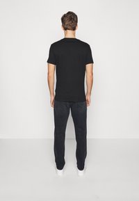 Camiseta de manga corta negra, material de algodón, cuello redondo. Combinada con jeans de mezclilla oscuros y zapatillas blancas, persona vista desde atrás.