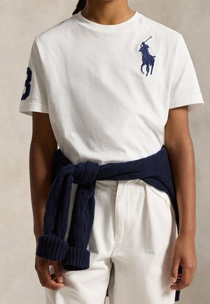 Personne portant un polo blanc avec un logo joueur de polo bleu marine et le numéro 3 sur la manche, pull bleu marine noué autour de la taille.
