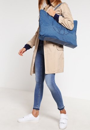 Modrá plátěná taška s velkým kruhovým logem, obsahující dva popruhy pro nošení a zipový uzávěr. Nesena přes rameno na světlém pozadí.