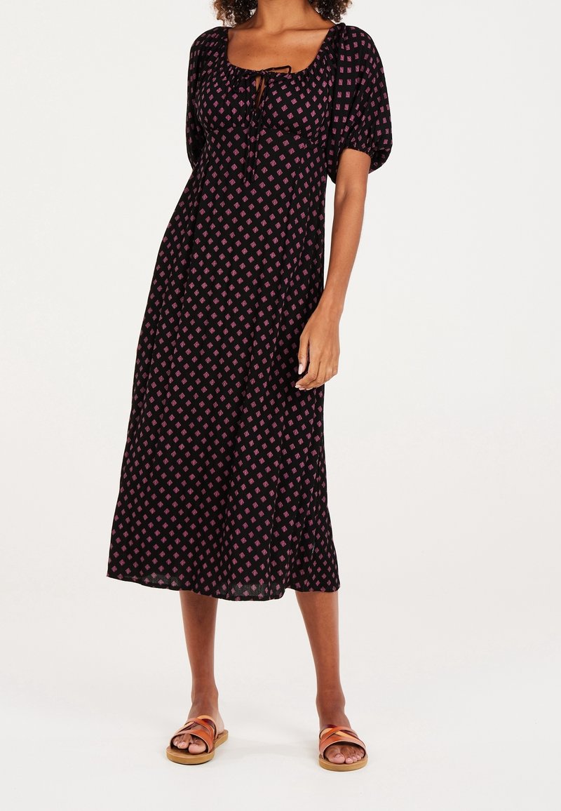 Robe noire avec motif de diamants roses, manches courtes bouffantes, encolure carrée, nouée à l'avant, et silhouette évasée en A. Portée avec des sandales plates.