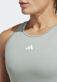 Top deportivo de color gris claro con una textura suave, que presenta un logo blanco en el lado izquierdo del pecho y tirantes delgados.