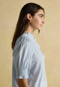 Frau mit langen braunen Haaren, die goldene Creolen trägt und eine hellblaue blusige Bluse mit Rüschen anhat, die nach rechts schaut, vor einem schlichten beigen Hintergrund.