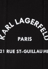 Crni materijal s podebljanim bijelim tekstom: "KARL LAGERFELD," "PARIZ," i "21 RUE ST-GUILLAUME," raspoređenim u kružnom dizajnu.