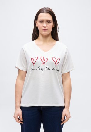 Hosszú sötét hajú nő, fehér V-nyakú pólót visel, amelyen három piros szív és kétszer ismétlődő "love always" felirat szerepel, sötét farmerrel párosítva.