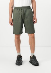 Mann i olivengrønne kne-lange shorts, hvite sokker og svarte joggesko, står mot en ensfarget hvit bakgrunn.