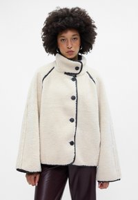 Crème-kleurige fleece jas met een hoge kraag, zwarte piping en vijf knopen. Heeft brede mouwen en een gestructureerde oppervlakte.