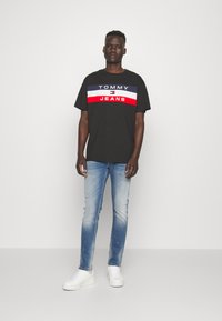 Čierne tričko s bielymi, námorníckymi a červenými horizontálnymi pruhmi s nápisom "TOMMY JEANS," spárované s svetlomodrými džínsami a bielymi teniskami.
