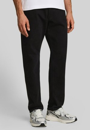 Mann trägt schwarze Hose und weiße Sportschuhe, steht vor grauem Hintergrund.