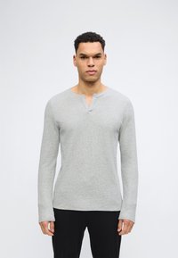 Άνδρας που φοράει ανοιχτό γκρι μακρυμάνικο πλεκτό μπλουζάκι με λαιμόκοψη σε σχήμα V και μαύρο παντελόνι, στέκεται μπροστά σε απλό λευκό φόντο.