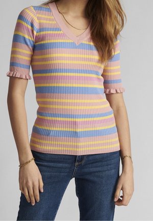 Kvinde iført en ribbet V-hals-top med pink, blå, gul og lilla vandrette striber og korte flæseærmer, parret med blå jeans.