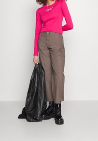 Žena oblečená v jasně růžovém topu s dlouhým rukávem, hnědých cargo kalhotách a černých bojových botách, drží černou koženou bundu v jedné ruce.