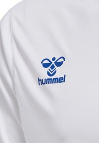 Weißer Stoff mit einem blauen, stilisierten Hummel-Logo und dem Text „hummel“, der auf dem Brustbereich eines Kleidungsstücks aufgedruckt ist.